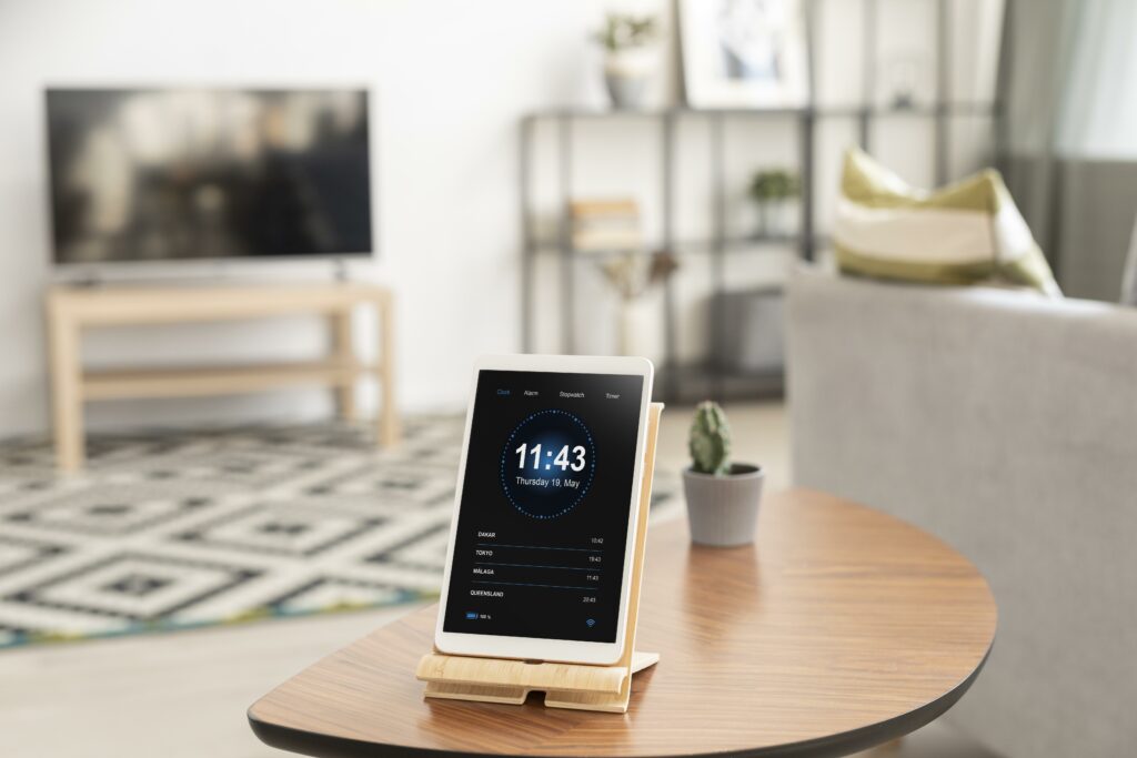 Modernes Wohnzimmer mit Tablet auf Holztisch, zeigt Weltzeituhr. Kaktus und Fernseher im Hintergrund.